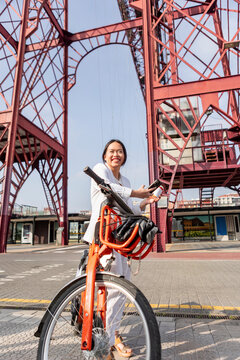 Smiling woman with bicycle beneath industrial steel structure