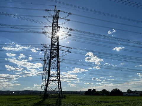 Silhouette of steel electricity pylon with power lines and sun shining through against blue sky with clouds