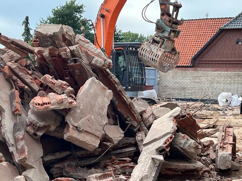 Pile of concrete and brick demolition rubble with excavator grapple in front of red brick house
