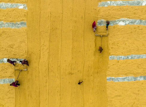 Ashuganj, Chittagong Division, Bangladesh - 05 July 2022: Aerial view of workers in colorful clothing raking and spreading golden rice grains on a large flat surface.