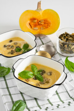 Two bowls of pumpkin soup with seeds and basil leaves garnished on white background