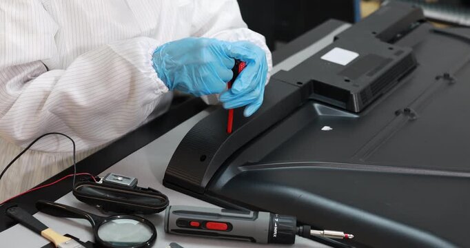 Technician in protective gloves repairs the back of a flat screen monitor with a screwdriver, using test tools on an electronics workbench.