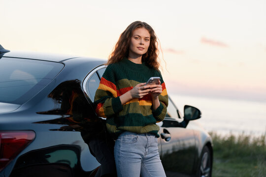 Young woman with curly hair in colorful sweater uses smartphone while leaning on black car by the sea at sunset. Travel and lifestyle concept.