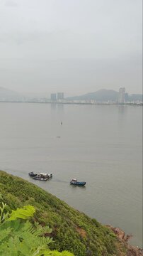 Aerial panoramic view of fishing boats sailing in the foreground with Quy Nhon city skyline in the background, Vietnam. Shot from Hai Minh peninsula featuring calm sea, coastal bay, and cityscape.