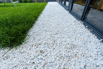 Fototapeta premium Strip of white decorative gravel between dark glass patio doors and lush green lawn in modern garden landscaping walkway