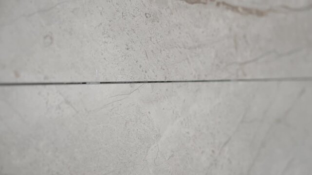 A slow, smooth tracking shot along the clean, dark grout line between two large, light grey marble-effect tiles. The camera reveals the subtle texture and veining of the modern, elegant surface