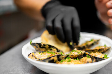 Chef in black glove arranging cooked mussels with a creamy sauce and parsley in a white bowl. © pavel siamionov