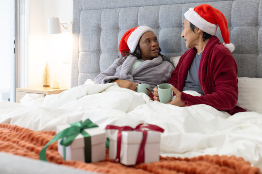 Couple Senior man  and  African American woman sitting in bed wearing Santa hats holding mugs christ