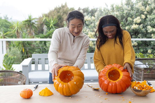 Senior male and adult female friends carving pumpkins on covered deck for halloween with tools