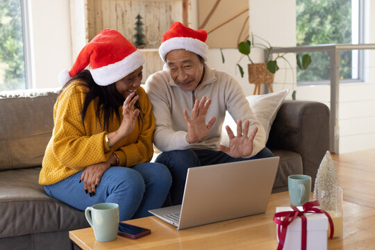 Senior Asian man, Adult African woman waving on sofa wearing Santa hats with laptop for christmas