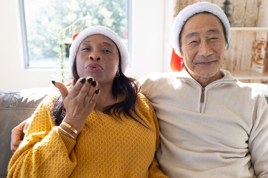 Senior Asian man  and  African-American woman sitting on sofa wearing Santa hats blowing kiss christ