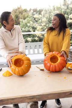 Diverse friends cleaning hollow halloween pumpkins on patio table with scooping spoon and seeds