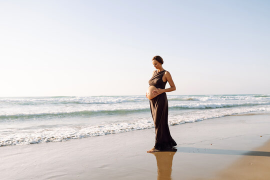 Pregnant woman walks along sunny beach by the water, touching her belly. Young woman in third trimester enjoys seaside scenery outdoors. Concept of enjoyment and freedom.