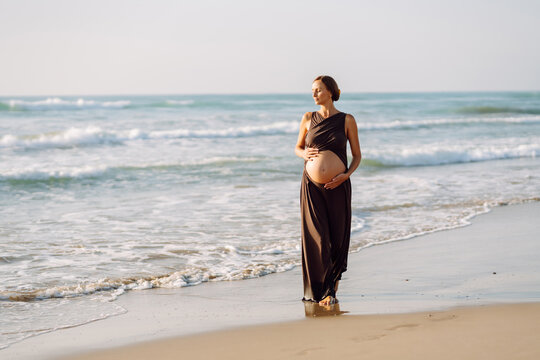 Pregnant woman walks along sunny beach by the water, touching her belly. Young woman in third trimester enjoys seaside scenery outdoors. Concept of enjoyment and freedom.