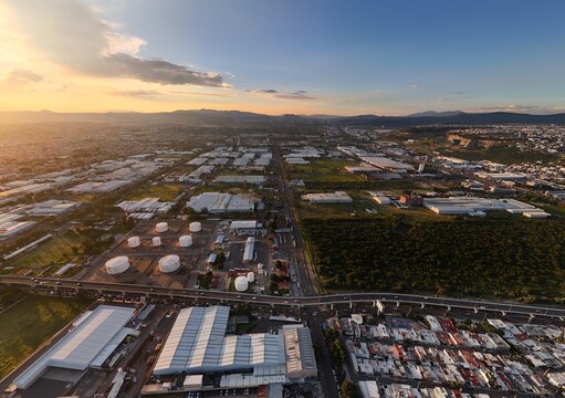 Santiago de Queretaro, Mexico - 18 September 2026: Aerial view of Parque Industrial Benito Juarez with industrial warehouses, storage tanks, and urban infrastructure under a golden sunset.