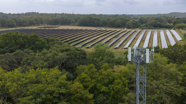 Fleet, England, United Kingdom - 18 September 2026: Aerial view of a solar farm with rows of panels, a mobile phone mast, and surrounding woodland under a cloudy sky.