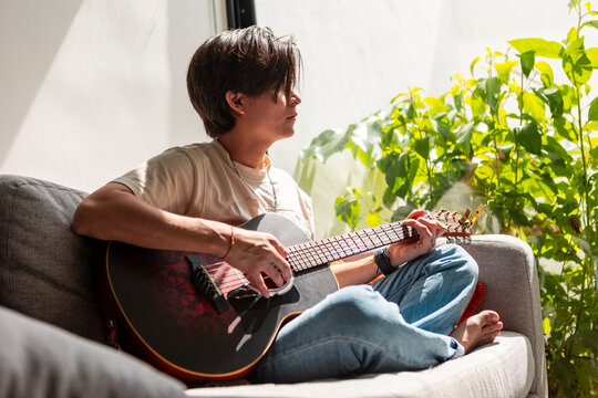 Young musician play acoustic guitar beside sunlit houseplant