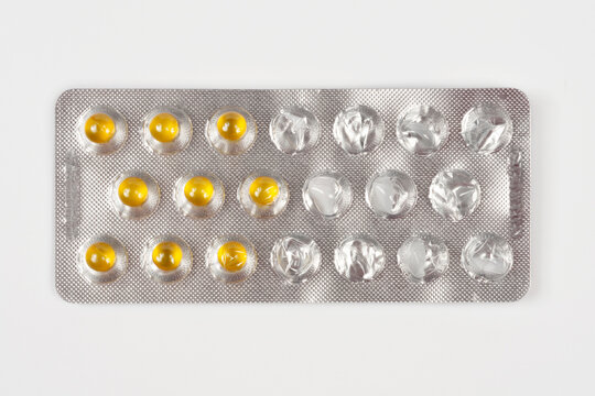 A top view of a partially used silver foil blister package containing nine remaining yellow softgel capsules and several empty slots over a solid grey background.