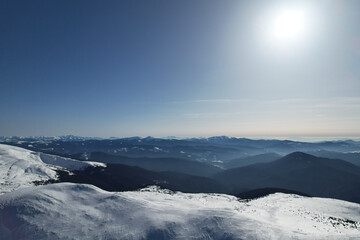 Obraz premium Aerial panoramic view of majestic high mountain peaks covered in snow under bright sun, drone shot of winter mountain ranges stretching to the horizon, sunny day in wintertime wilderness, blue sky