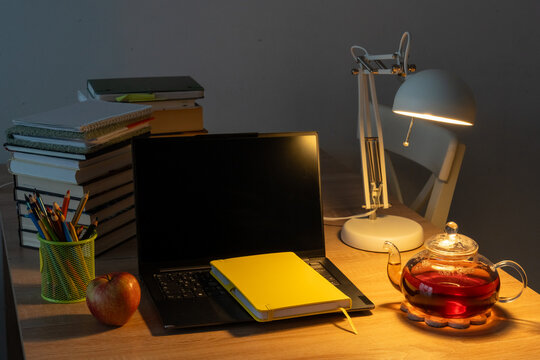 Glass teapot of brewed tea beside laptop, yellow notebook, books and apple on a warmly lit desk. Concept of quiet evening study, mindful work breaks and a cozy home learning routine.