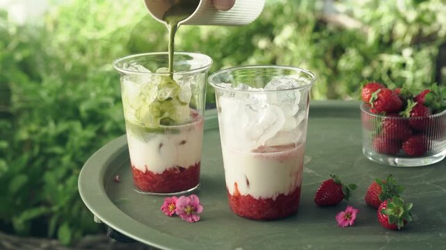 Close-up of green matcha being poured over ice and milk to create a layered strawberry matcha latte. Fresh strawberries and flowers decorate the outdoor table in a lush summer garden setting