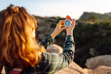 Fototapeta premium Female hiker with backpack holds compass while standing on mountain on sunny day. Young woman enjoys adventure and looks at compass at sunset. Concept of nature, navigation, and travel.