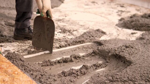 Close up scene shows worker shaping wet concrete across rough foundation surface with trowel, surrounded by boots, gloves, wooden formwork, and soft warm outdoor light in unfinished building area.