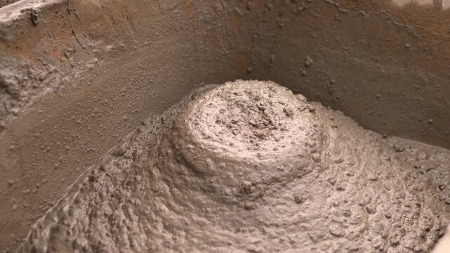 Close up of thick wet concrete or mortar inside container, where small clump drops into rounded mound, leaving rough swirl marks and muddy granular walls under soft natural light.
