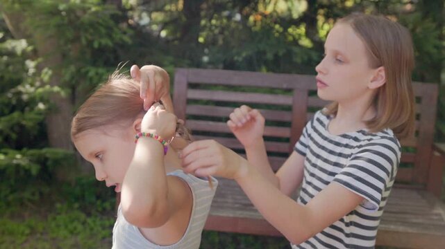 Older sister gently styles child hair outside on wooden seat in backyard. Happy siblings enjoy peaceful moments braiding locks together with joyful connection under bright natural daylight.