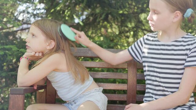 Young bored child outdoor looks away while sister combs straight blonde hair on wooden park bench. Little girl displays indifferent emotion during simple grooming process under sunlight.