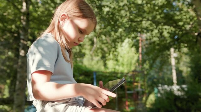 Child taps screen on digital tablet during outdoor homeschooling session under garden trees. Focused young girl participates in remote education online using portable modern device outside.