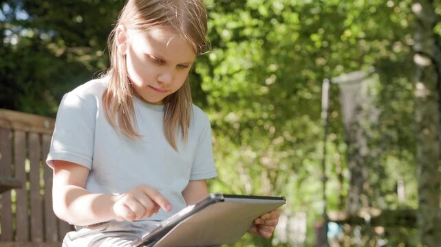 Attentive child taps digital tablet computer outside in backyard garden. Schoolchild engages in outdoor learning activity using touchscreen device during bright sunny summer afternoon.