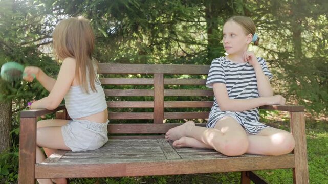 Two girls sit outdoors on wooden bench during child hair grooming routine, holding hairbrush playfully in backyard garden environment under bright natural sunlight with relaxed mood outdoors.