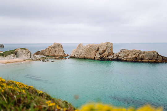 A view of a sandy beach with rock formations and shallow water. Gray clouds are visible in the sky. Signs of the receding tide are evident along the shoreline.