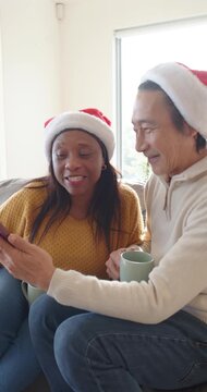 Vertical video: Diverse couple senior Santa hats showing phone on couch laughing at christmas mugs