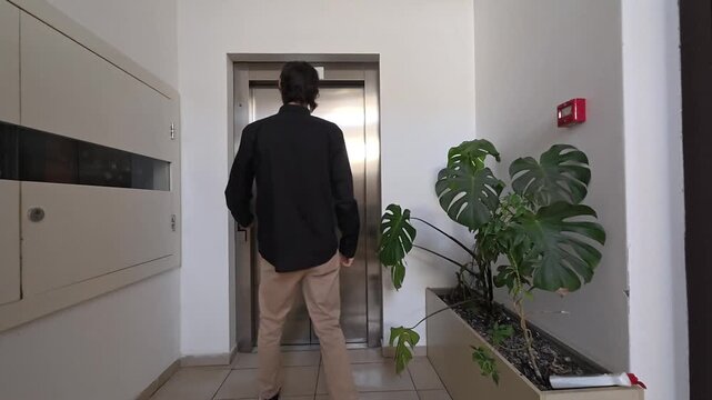 Man waits alone for elevator in quiet building hallway, then enters as doors open. Elevator passenger stands patiently beside leafy indoor plant, conveying solitude during everyday transit.