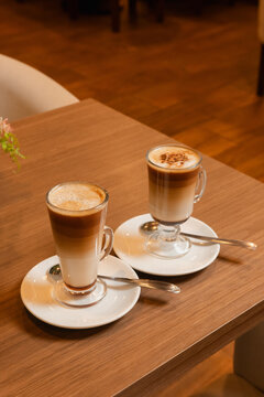 Two cups of cappuccino in tall glass mugs on white saucers with small spoons.