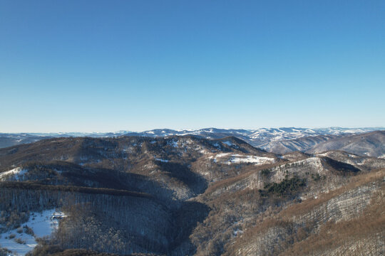 Aerial view of winter mountains covered in forest and patches of snow under bright blue sky, drone panoramic shot of scenic mountain ranges and deep valleys, sunny wintertime landscape, carpathians