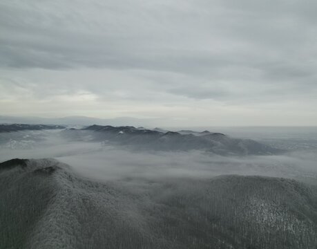 Aerial view of winter mountains covered in frozen forest with dense fog rolling through valleys, drone shot of snowy ridges under gray cloudy sky, moody wintertime wilderness landscape, misty weather