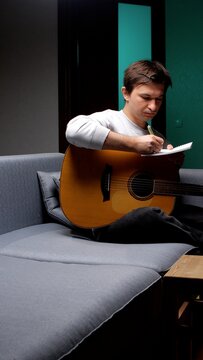 Young man on a sofa with an acoustic guitar, jotting lyrics and chords in a notebook while composing music at home, focused on melody, creativity and songwriting practice