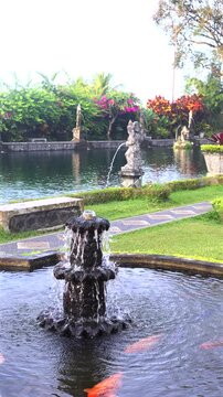 Ornamental fountain pours into koi pond as orange fish glide beneath surface. Stone sculpture, stone path, and vivid shrubs create tranquil garden scenery with reflective water and soft motion.