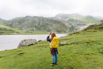 Obraz premium Female hiker with a yellow backpack uses her phone to photograph a mountain-ringed lake. She is enjoying nature and holding her smartphone on a cloudy day. Nature and adventure concept.