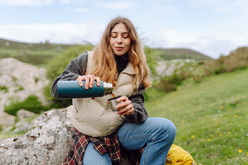 Naklejka premium Female hiker with backpack sits on large rock, pouring tea from thermos against picturesque mountain backdrop. Woman enjoys a rest and drinks a hot beverage while hiking. Adventure concepts.