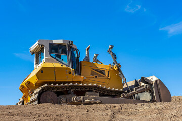 Massive, bright yellow bulldozer at construction site with tracks embedded in a mound of rich, brown soil. A clear blue sky creating a contrasty background. © beachfront