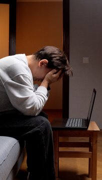 Man feeling frustrated and depressed, covering his face with hands while sitting on a sofa with a laptop, representing overwhelming pressure, emotional exhaustion, and mental health issues
