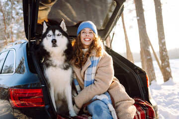 Woman and her husky are relaxing in trunk of car in snowy forest. Happy woman enjoys winter outing with dog, sitting on blanket in car at sunset. Concept of friendship and adventure. Lifestyle. © maxbelchenko