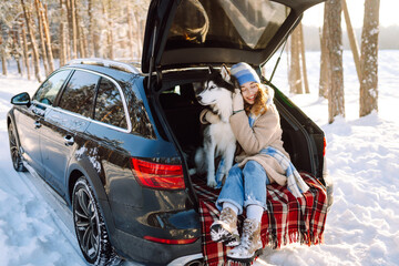 Woman and her husky are relaxing in trunk of car in snowy forest. Happy woman enjoys winter outing with dog, sitting on blanket in car at sunset. Concept of friendship and adventure. Lifestyle. © maxbelchenko