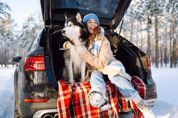 Woman and her husky are relaxing in trunk of car in snowy forest. Happy woman enjoys winter outing with dog, sitting on blanket in car at sunset. Concept of friendship and adventure. Lifestyle. © maxbelchenko