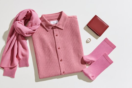 Pink mens clothing flat lay with textured button shirt, draped scarf, folded socks, red wallet and ring arranged on a light background, overhead fashion still life