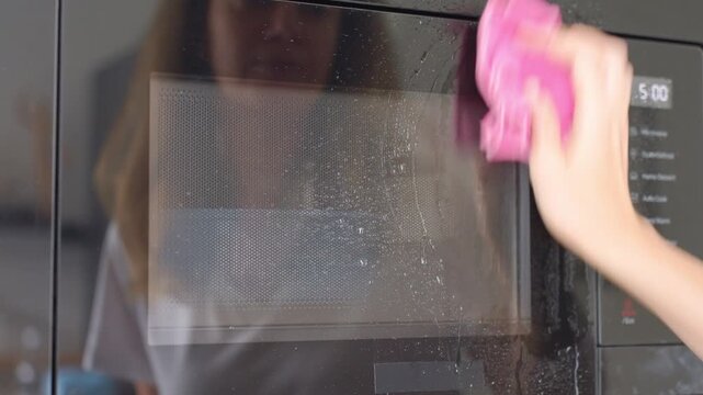 Hand sprays cleaner across microwave glass door and wipes wet surface with pink cloth. Concept of appliance cleaning, kitchen hygiene and household maintenance